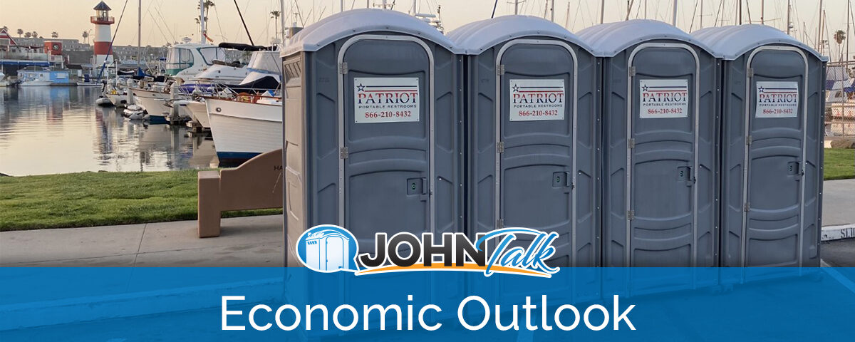 Gray portable toilets next to water with boats and a lighthouse in the background