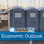 Gray portable toilets next to water with boats and a lighthouse in the background