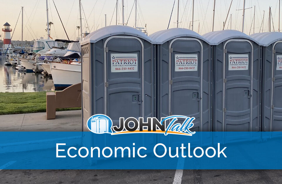 Gray portable toilets next to water with boats and a lighthouse in the background