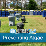 A group of portable sinks and trash cans in a grassy field with a row of blue and yellow portable toilets in the background