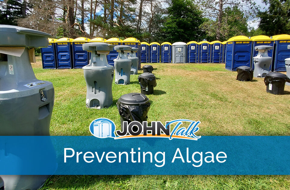 A group of portable sinks and trash cans in a grassy field with a row of blue and yellow portable toilets in the background