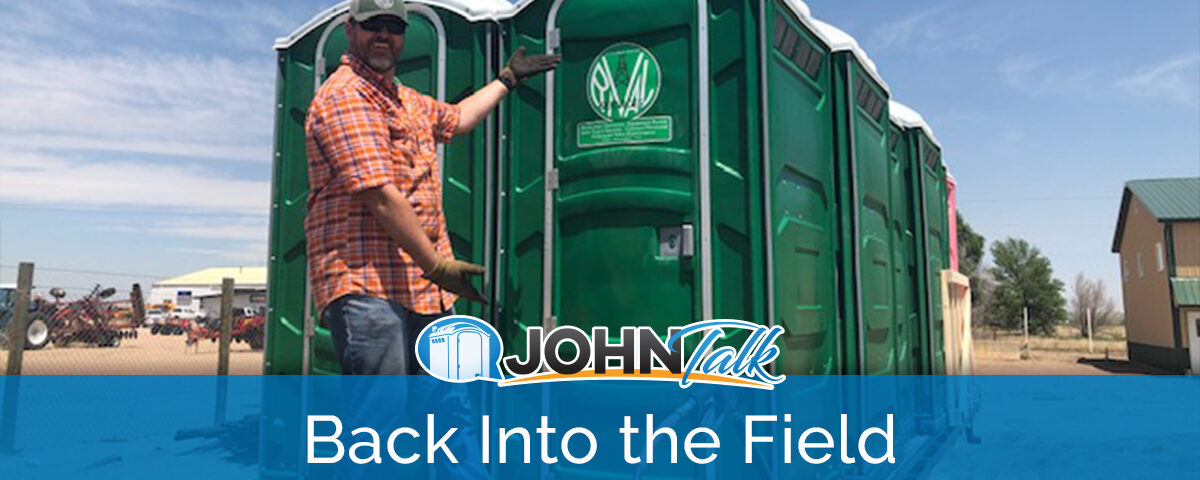 A man in an orange shirt, cap and sunglasses, wearing gloves, motioning to a group of green portable toilets on the back of a flatbed trailer