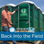 A man in an orange shirt, cap and sunglasses, wearing gloves, motioning to a group of green portable toilets on the back of a flatbed trailer