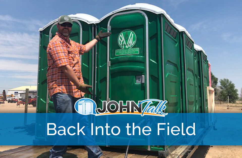 A man in an orange shirt, cap and sunglasses, wearing gloves, motioning to a group of green portable toilets on the back of a flatbed trailer